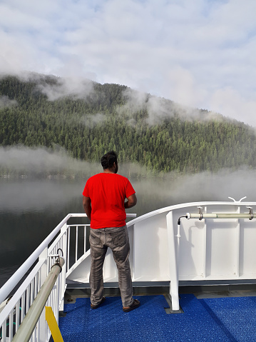 Klemtu, Canada - May 17, 2020. A man looks at the foggy, mountainous shoreline on deck of a ferry as it departs from Klemtu in Coastal BC.