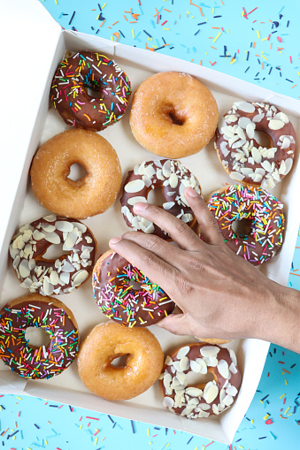 Stock photo showing close-up, elevated view of rows of alternating plain and chocolate glazed ring doughnuts in cardboard box. Some of the chocolate glazed cakes are topped with flaked almonds, whilst others are decorated with white, pink, green, blue, yellow, and orange, hundred and thousand sugar sprinkles.