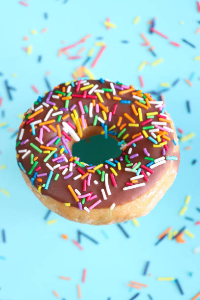 Stock photo showing close-up, elevated view of individual chocolate glazed ring doughnut decorated with white, pink, green, blue, yellow, and orange, hundred and thousand sugar sprinkles on blue background.