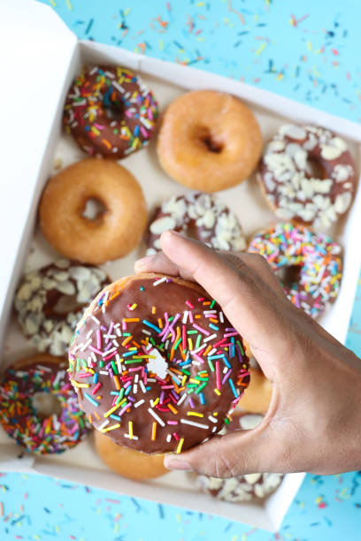 istock Close-up image of unrecognisable person holding glazed ring doughnut from cardboard box, plain and chocolate glazed fried dough dessert decorated with multicoloured sugar sprinkles or flaked almond nuts, blue background, elevated view 1873838358