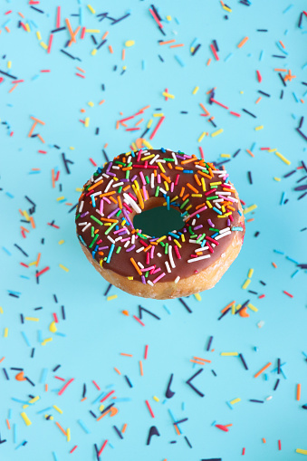 Stock photo showing close-up, elevated view of individual chocolate glazed ring doughnut decorated with white, pink, green, blue, yellow, and orange, hundred and thousand sugar sprinkles on blue background.