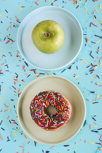 Stock photo showing close-up, elevated view of two plates, one containing a chocolate glazed ring doughnut decorated with white, pink, green, blue, yellow, and orange, hundred and thousand sugar sprinkles, the other with a green apple. Food choices concept.