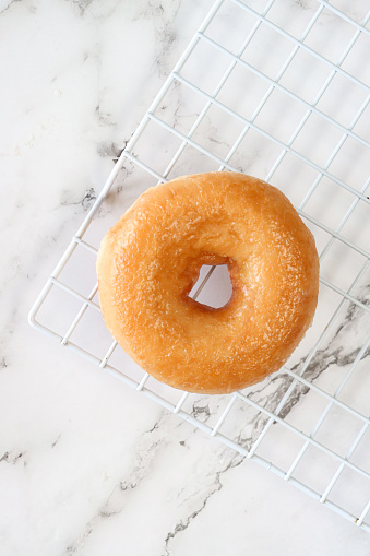 Stock photo showing close-up, elevated view of a glazed ring doughnut on white cooling rack over marble effect background.