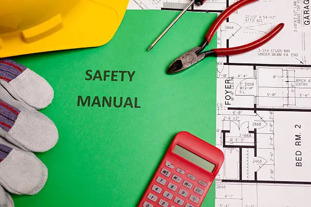 A safety manual is surrounded by work gloves, a hardhat, calculator, work tools, and floor plans.