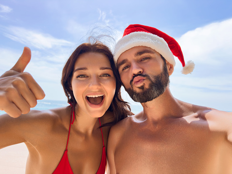 Christmas couple selfie photo during beach vacation at winter holidays. Happy young adults smiling at camera taking self-portrait with thumb up wearing Santa hat. Multiracial Caucasian and Asian people. High quality photo