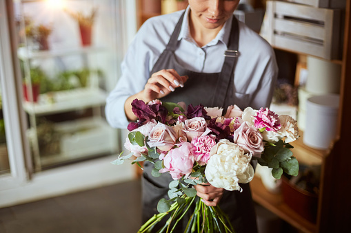 Portrait of female florist in apron arranging fresh flowers for bouquet in the flower shop, using roses, hydrangea, peonies