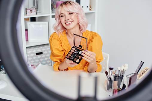 Young beautiful blond asian woman is recording make up tutorial video for beauty blog, wearing terracota blouse. Video blogging, isolation, stay home concept