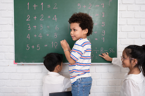 Happy African American kid boy studying and writing with chalkboard and friend in classroom
