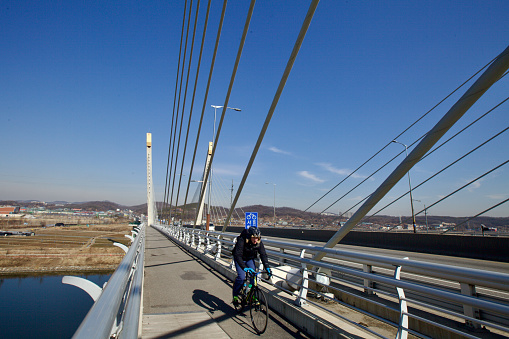 Incheon, South Korea - February 10, 2020: Cyclists traverse the Baekseok Bridge, its cables creating a striking pattern above the serene Gyeongin Ara Waterway.