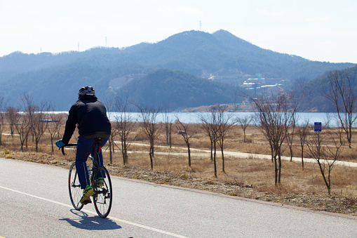 Yeoju City, South Korea - February 20, 2020: A lone cyclist braves the winter chill on the Hangang Bike Path, with barren trees and mountain backdrop along the South Han River.