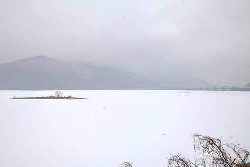 Namyangju City, South Korea - February 15, 2020: Overcast skies loom over the frozen expanse of Paldang Lake, scattered with snow-covered islets and ice, hinting at nature's resilience.
