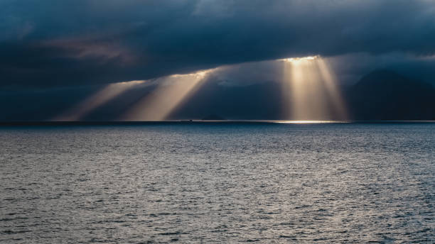 At sunset, the sky is covered with dense clouds. The sun's rays filter through the clouds, casting a soft glow on the surface of the sea. Rows of mountains silhouette the distant landscape. The densely clouded sky creates a dramatic atmosphere, and the sunlight reflecting on the sea adds a mystical touch to the scene. Nature's enchantment embellishes this magnificent moment.