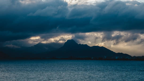 At sunset, the sky is covered with dense clouds. The sun's rays filter through the clouds, casting a soft glow on the surface of the sea. Rows of mountains silhouette the distant landscape. The densely clouded sky creates a dramatic atmosphere, and the sunlight reflecting on the sea adds a mystical touch to the scene. Nature's enchantment embellishes this magnificent moment.