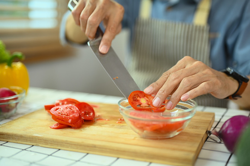 Senior man cooking with organic food in kitchen, slicing ripe tomato on wooden board