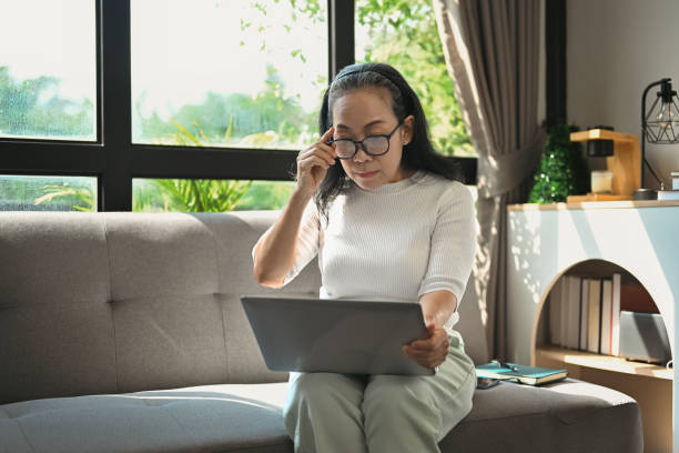 istock Middle age woman touching glasses and reading email on laptop while relaxing on the couch at home. 1874143046