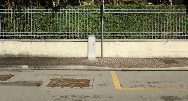 istock Fence made of low white plaster wall with high sharped railing on top. Old utility box in middle. Hedge on behind , sidewalk and city street in front. 1874143361