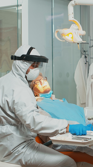 Dentistry doctor in protective suit using sterilized dental tools examining senior patient during coronavirus pandemic. Medical team talking with woman wearing face shield, coverall, mask and gloves