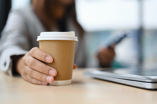 Closeup female worker hand holding a paper cup of hot coffee and using mobile phone