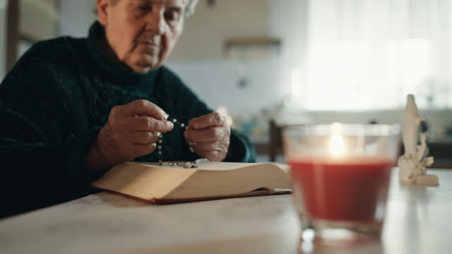 SLO MO Rack Focus Shot of Senior Woman with Rosary Beads and Holy Bible at Table with Lit Candle at Home