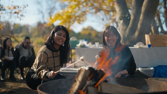 Two multi-ethnic women friends are enjoying eating marshmallows in nature in a camping site.