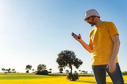 Castilla La Mancha, Spain. Low angle view of a bearded mature man with a sun hat, looking at his mobile phone in a flowery field on a clear and sunny spring afternoon.