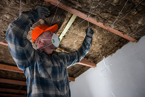 a worker in overalls, gloves and a respirator attaches the glass wool to the ceiling with a cord