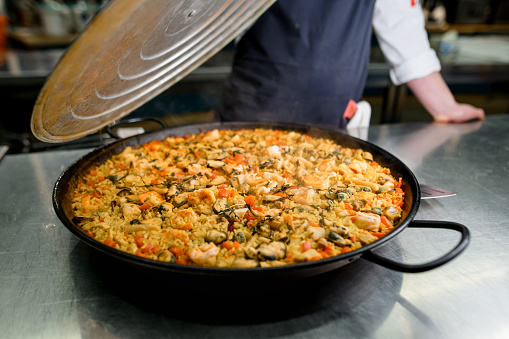 Chef removes the lid from the pan and presents traditional Paella with shrimp, mussels and vegetables.
