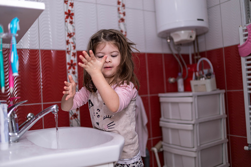The cute, three-year-old girl is standing by the sink and looking at her arm to see where the water drop is going.