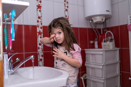 Adorable little girl is standing by the sink and using running water to wash her eyes.