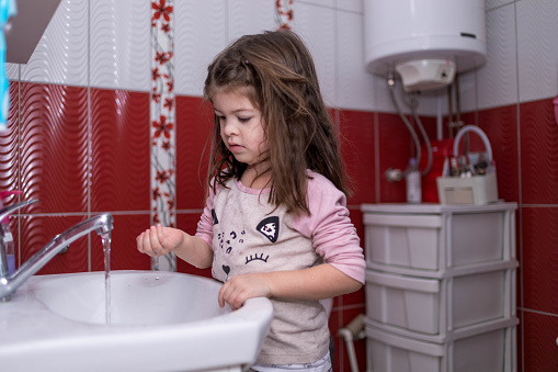 The child is playing with running water in the bathroom. The toddler is standing by the sink , and washing her hands and face .