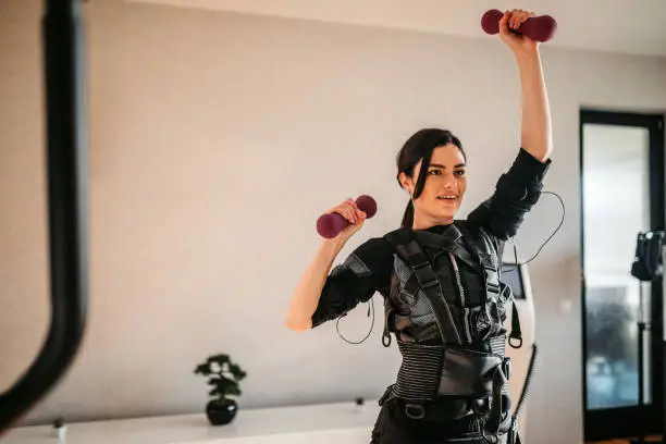 Beautiful athletic woman wearing an EMS suit while training with dumbbells in the gym.