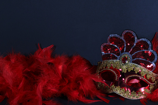 Red and gold carnival mask on an illuminated black background, placed on the right, above a garland of red feathers, in the middle distance. With copy space