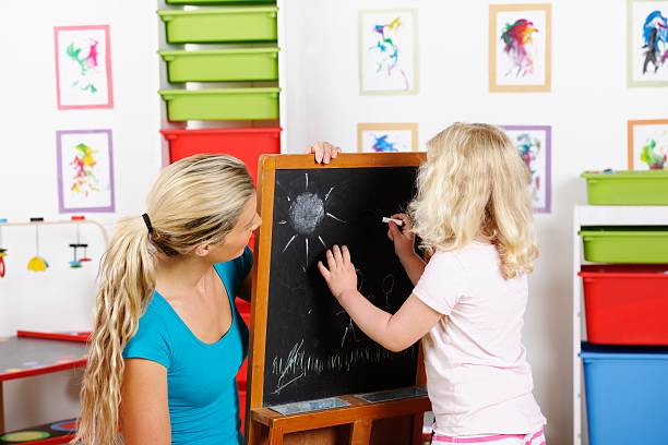 istock Little Girl Being Supervised Whilst Writing On The Blackboard 187448254