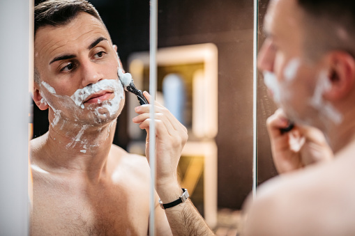 Handsome young man shaving his beard while looking in the bathroom mirror.
