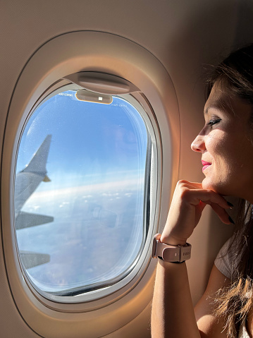 Young woman looking happily through the airplane window. Traveling girl enjoying the view of the sky and clouds in mid-flight.