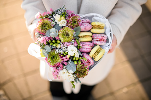Top view on heart shaped box with mix of fresh flowers and colorful french macarons in woman's hands. Present for holiday. Gift shop