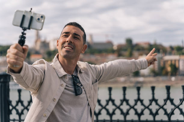 istock Young content creator filming himself with his cell phone on a tripod while talking to his followers. Boy filming with his cell phone pointing the river and the city of Budapest. 1874539010