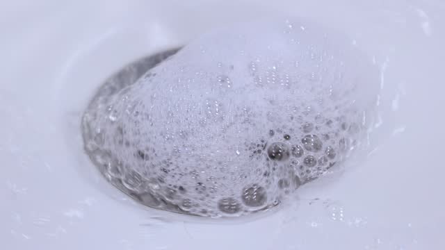 Metallic drain hole in the ceramic sink with flowing water with white soap foam in the bathroom close up