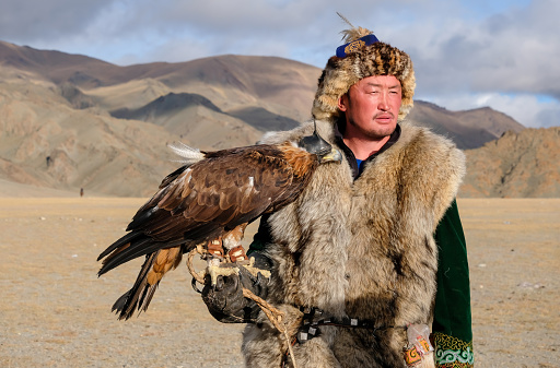 A Kazakh eagle hunter standing with his hooded eagle at the Sagsai Golden Eagle Festival held on 17th-18th September 2023, near the small town of Bayan-Ulgii province district of Western Mongolia. Set in the Altai Mountain range, the nomadic eagle hunters, dressed in traditional fur and embroidered clothing, gather to compete with their eagles, in various competitions that show the skill and bond between the hunter and their eagle.