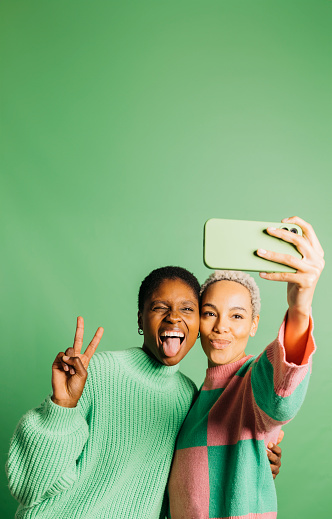 Two young happy women taking a selfie in front of a green background in a studio. Women taking a selfie in a studio.