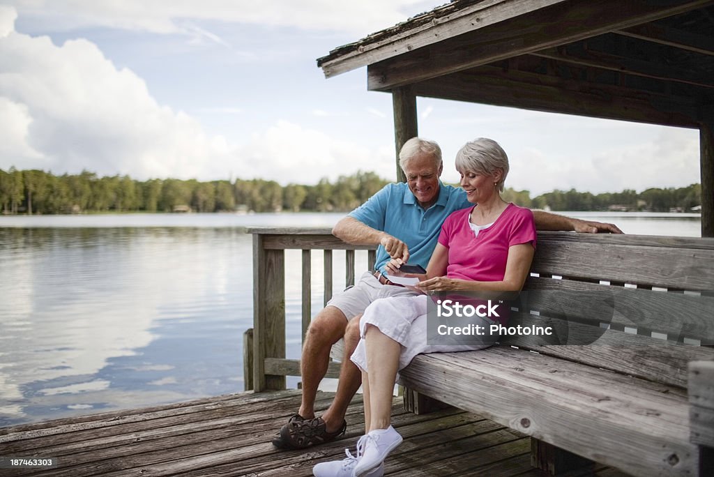 Senior Couple Using Mobile Phone To Deposit Bank Check Happy senior couple using mobile phone to deposit bank check on wooden bench near pond Bank Deposit Slip Stock Photo