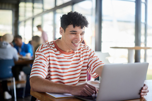 Young male college student laughing while doing his homework on a laptop at a table in a campus cafeteria