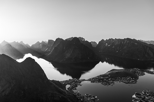 A monochrome view of Reinebringen reveals the stark contrasts between the rugged mountains of the Lofoten Islands and the reflective waters below, with the silhouette of Reine village etched into the landscape (Vintage Effect)