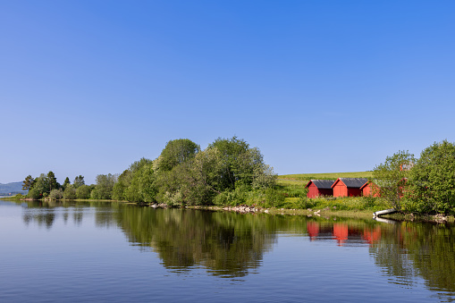 A vibrant summer scene at Snasavatnet, Norway, featuring charming red boat houses along the lake's shore against a backdrop of clear blue skies, epitomizing the picturesque and serene summer ambiance of the Steinkjer region