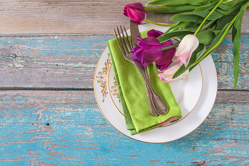 bouquet of purple and white tulips on the whte plates with green napkin and vintage fork and knife on a old blue paint wooden background; copy space