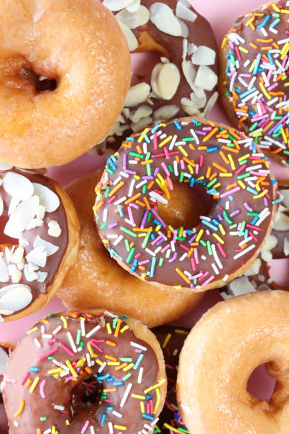 istock Full frame image of stacked mixed, glazed ring doughnuts, plain and chocolate glazed fried dough dessert decorated with multicoloured hundreds and thousands, sugar sprinkles or flaked almond nuts, pink background, elevated view 1874775712