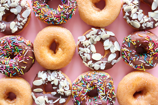 Stock photo showing close-up, elevated view of rows and columns of alternating plain and chocolate glazed ring doughnuts on pink background. Some of the chocolate glazed cakes are topped with flaked almonds, whilst others are decorated with white, pink, green, blue, yellow, and orange, hundred and thousand sugar sprinkles.