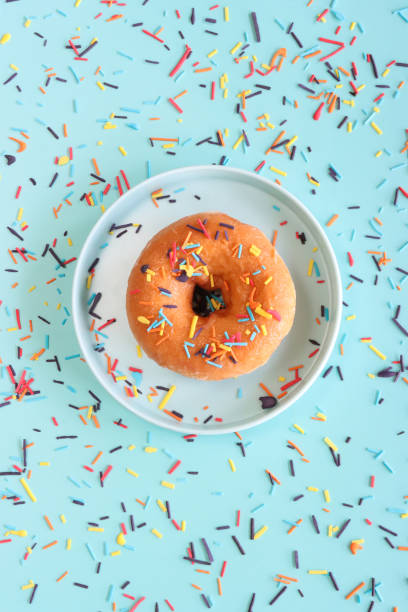Stock photo showing elevated view of a blue plate containing an individual glazed ring doughnut decorated with white, pink, green, blue, yellow, and orange, hundred and thousand sugar sprinkles on blue background.