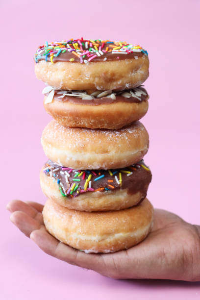 istock Image of unrecognisable person balancing stack of alternating plain and chocolate glazed ring doughnuts on palm of hand, fried dough dessert decorated with multicoloured sugar sprinkles or flaked almond nuts, pink background, focus on foreground 1874784602