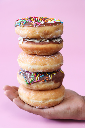 Stock photo showing close-up view of a stacked batch of alternating plain and chocolate glazed ring doughnuts balanced on the palm of an unrecognisable person's hand. Some of the chocolate glazed cakes are topped with flaked almonds, whilst others are decorated with white, pink, green, blue, yellow, and orange, hundred and thousand sugar sprinkles.